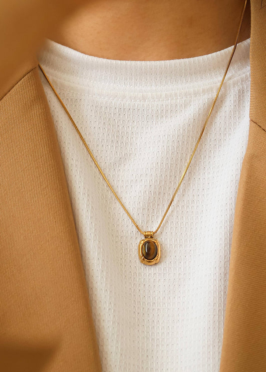 Gold necklace with a pendant worn by a person in a white top and brown jacket.