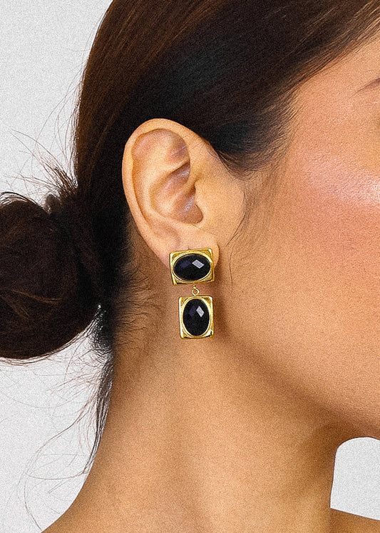 Close-up of a person wearing gold earrings with black stones on a neutral background