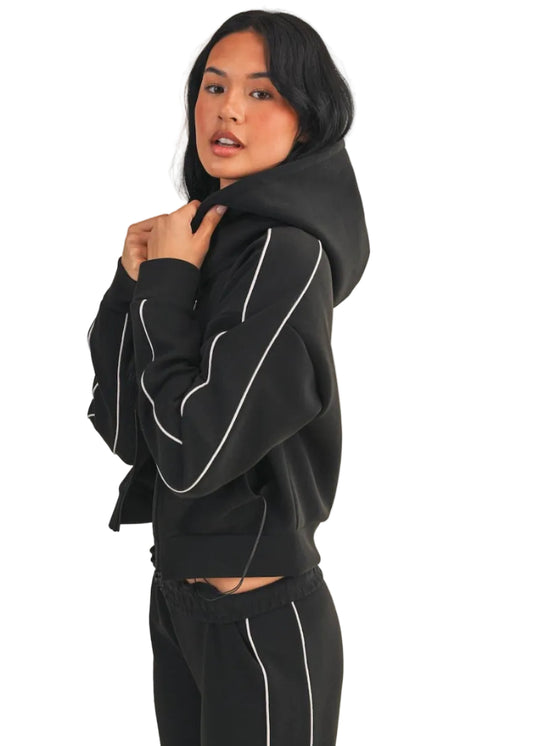 Woman wearing a black hoodie and pants with white piping on a beige background