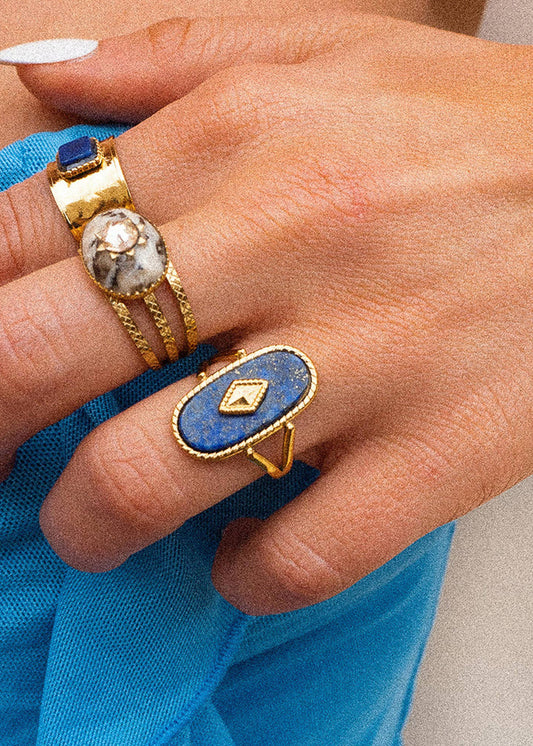 Close-up of a hand wearing two gold rings with blue stones against a blue fabric background