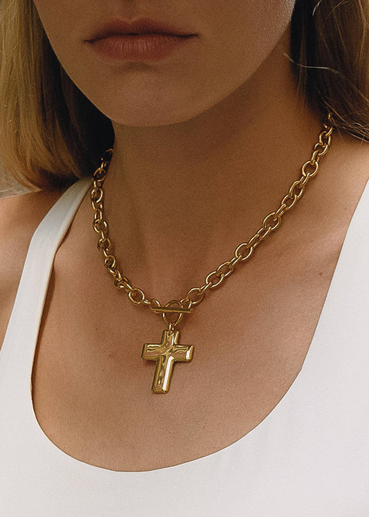 Gold chain necklace with a cross pendant worn by a person in a white top.