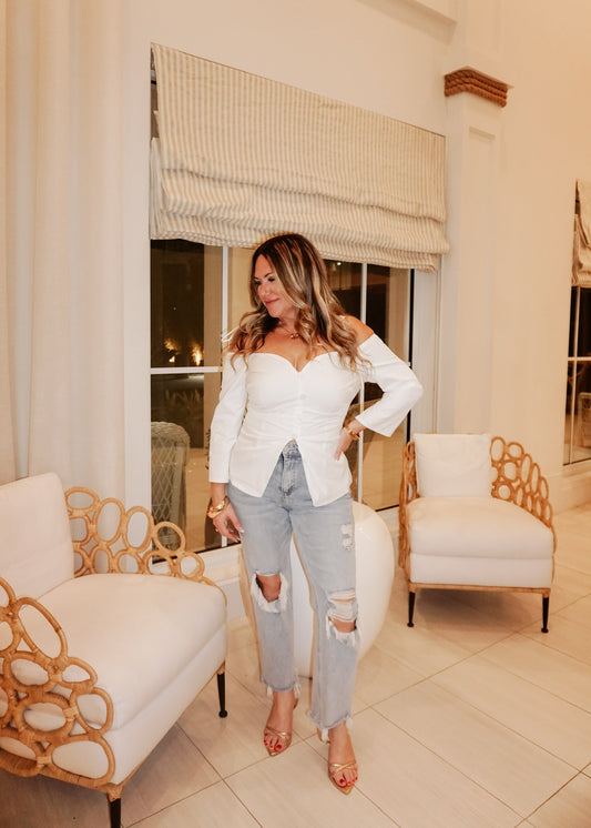 Woman in a white blouse and ripped jeans standing in a stylish room with decorative chairs and a window.