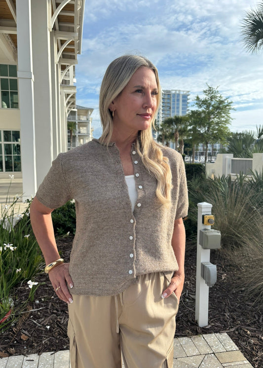 Woman in beige cardigan and pants standing outdoors with buildings and trees in the background