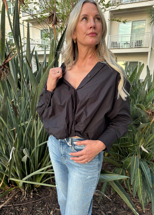 Woman wearing a dark shirt and light blue jeans standing outdoors with greenery and buildings in the background