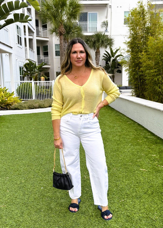 Woman in yellow cardigan and white pants standing on grass with a building and palm trees in the background