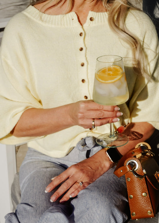 Person holding a glass of lemonade with a lemon slice, wearing a yellow cardigan and gray pants.