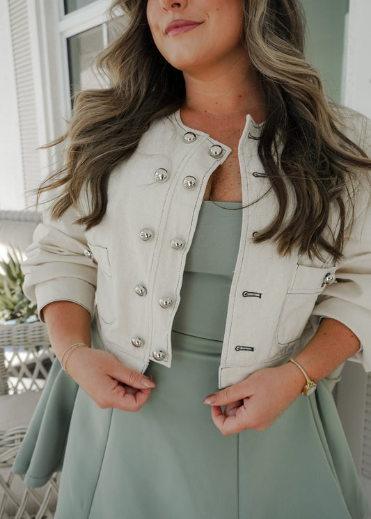 Woman wearing a cream jacket with silver buttons over a green dress.