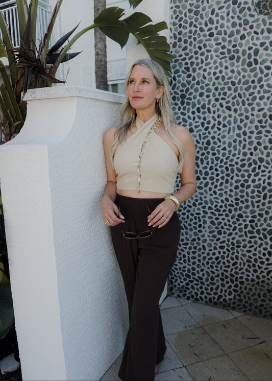 Woman in beige top and black pants standing against a textured wall with plants.