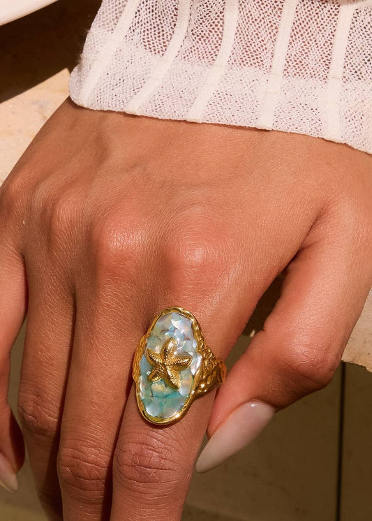 Hand wearing a gold ring with a large blue stone on a neutral background