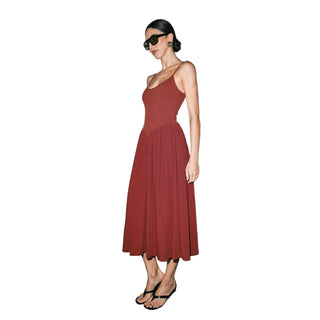A woman modeling a sleeveless, scoop-neck midi dress in a dusty rose or muted terracotta red. She is wearing black sunglasses, black strappy sandals, and holding a light beige shoulder bag, standing against a plain light gray wall.