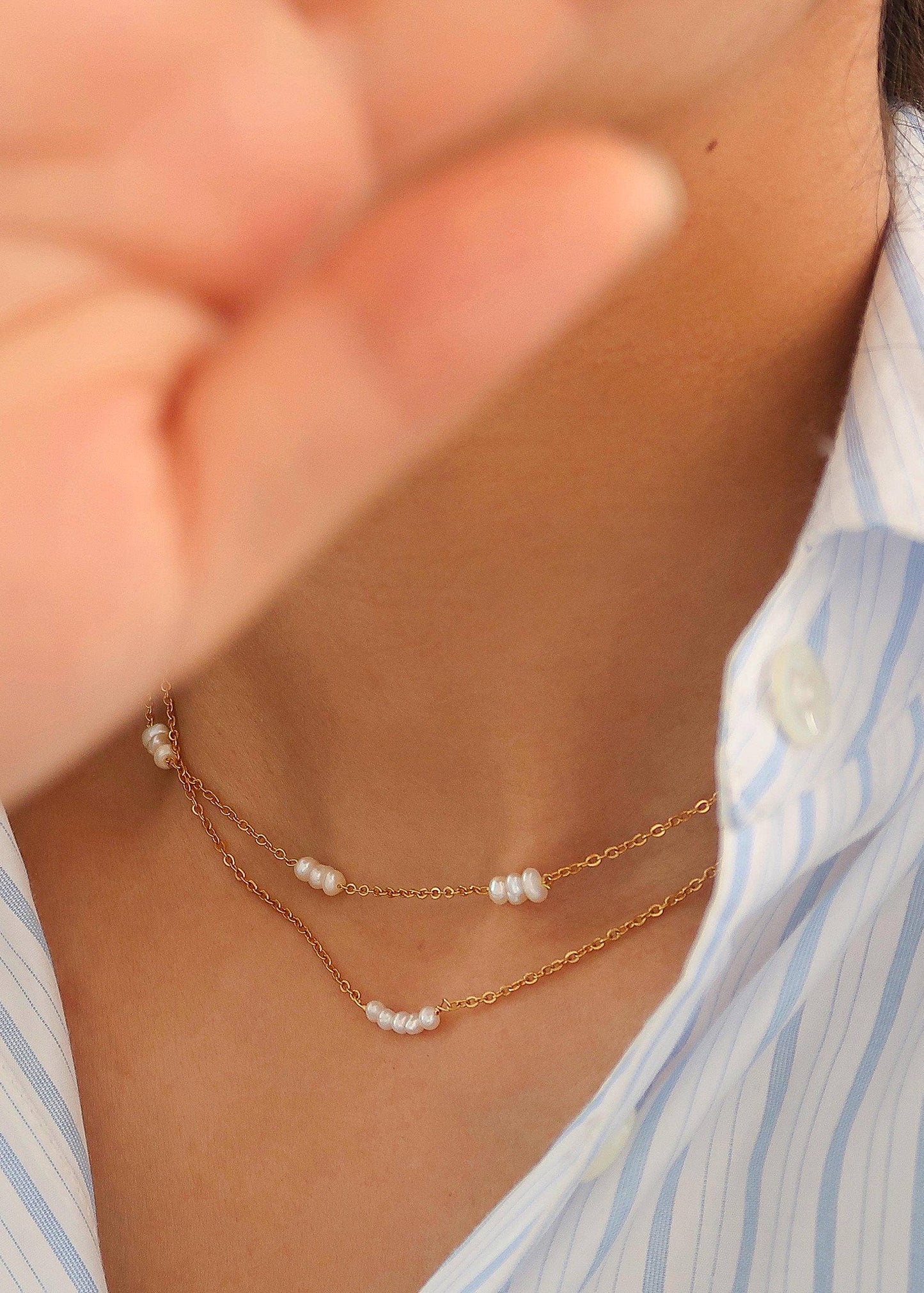 A close-up image of a woman wearing a petite pearl choker with 18K gold plating on stainless steel, displayed against a striped shirt background.