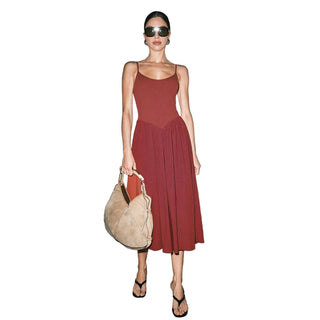 A woman modeling a sleeveless midi dress in a deep rust red color, paired with a beige suede hobo bag and black sandals, standing against a neutral light gray wall.