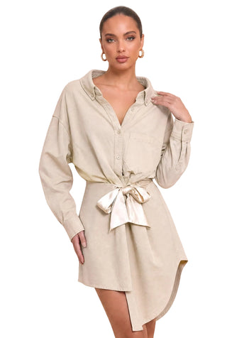 A woman models a light beige, oversized long-sleeve shirt dress with a tie waist and asymmetrical hem against a solid beige background.