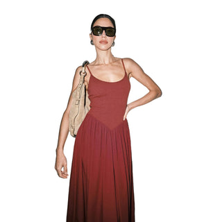 A woman modeling a rust-red spaghetti-strap maxi dress with a pleated skirt, styled with dark sunglasses and a beige shoulder bag against a plain light gray background.