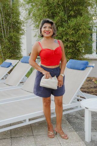 Woman in a red top and blue shorts standing on a patio with lounge chairs and greenery.