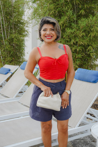Woman in a red top and blue shorts holding a white clutch by lounge chairs with greenery in the background