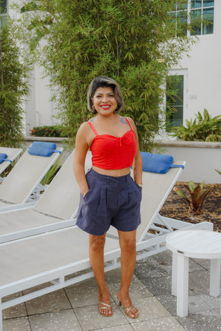 Woman in a red top and blue shorts standing in a sunlit outdoor area with lounge chairs and plants.