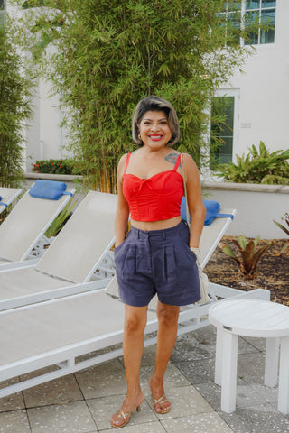 Woman in a red top and purple shorts standing in an outdoor setting with lounge chairs and plants.