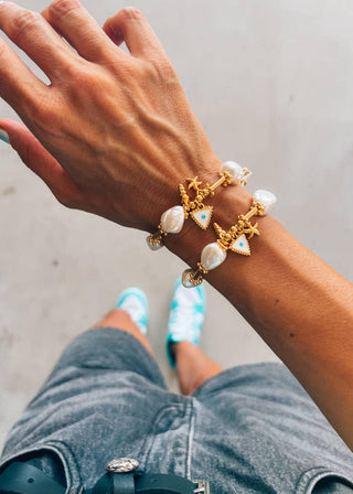 Hand wearing two gold bracelets with pearl-like stones on a neutral background.