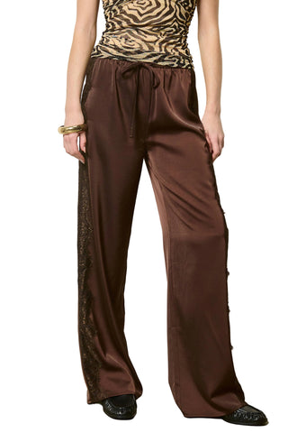 A model wearing wide-leg brown satin pants with black lace side panels and a drawstring waist, paired with a leopard-print top and black shoes, standing against a plain off-white wall.