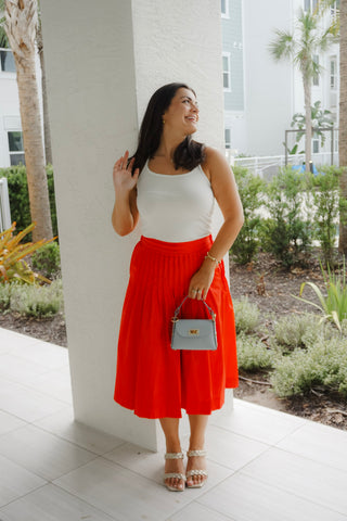 Woman in a white top and red skirt standing outdoors near a building.