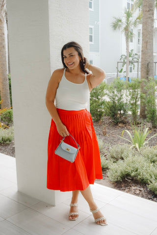 Woman in a white top and orange skirt standing outdoors near a building.