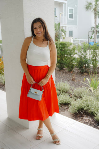 Woman wearing a white top and red skirt with a small handbag, standing on a patio.