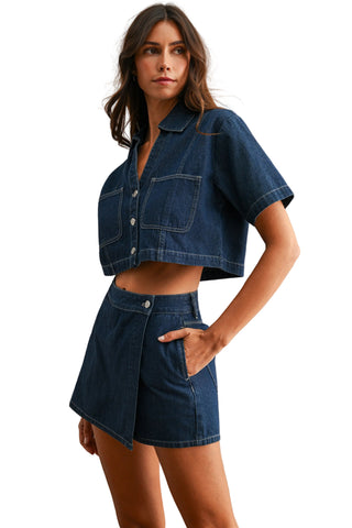 A woman modeling a dark blue denim two-piece set consisting of a short-sleeve cropped button-up shirt and a matching high-waisted wrap mini skirt, set against a neutral textured beige wall.