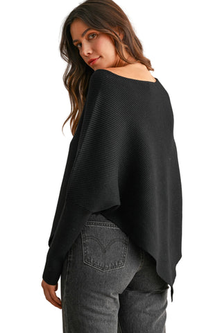 A woman wearing a black off-the-shoulder ribbed knit poncho sweater and gray jeans, photographed against a neutral beige studio background.