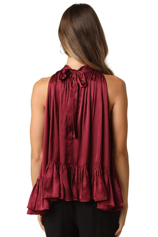 A woman shown from the back wearing a sleeveless deep burgundy satin top with a tie-neck detail and ruffled hem, paired with black pants, against a solid beige background.