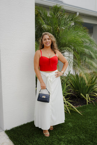Woman in a red top and white skirt standing outdoors with greenery in the background