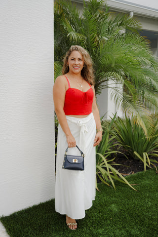 Woman in a red top and white pants standing outdoors with plants in the background