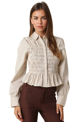 A woman modeling a cream and brown vertically striped long-sleeve blouse with a smocked bodice and peplum hem, paired with dark brown pants, against a solid beige background.