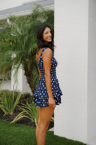 Woman in a blue polka dot dress standing next to a white wall with greenery.