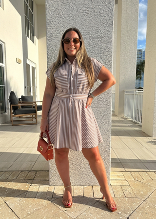 A woman wearing a sleeveless, short-sleeve button-up mini dress with vertical pink and white stripes, standing outdoors in front of a textured white column. She is holding a small reddish-brown handbag and wearing clear-strap high heels.