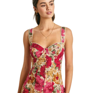 A woman wearing a sleeveless floral corset-style dress with pink and red flowers on a beige background, photographed against a light paneled wall.