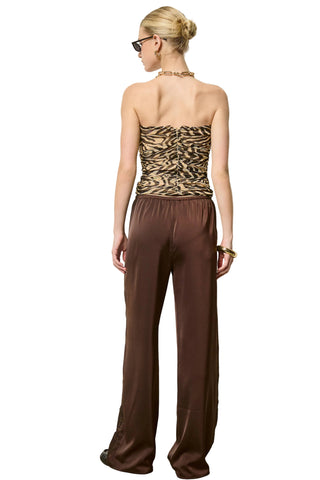 A woman stands with her back to the camera wearing a strapless tube top with a tan and black zebra-like pattern and matching wide-leg brown pants against a plain off-white wall.