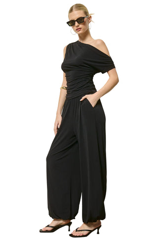 A woman modeling a black one-shoulder draped jumpsuit with wide legs, styled with black sunglasses and black sandals against a plain light gray wall.