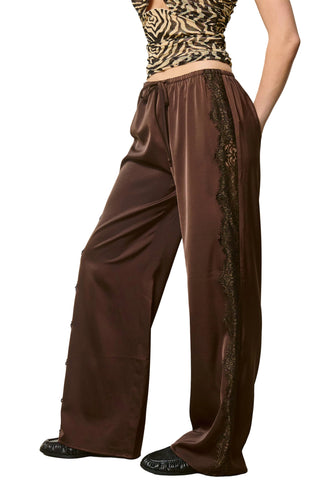 A person wearing wide-leg brown satin pants with black lace trim down the sides, paired with a patterned top and black shoes, standing against a plain light wall.