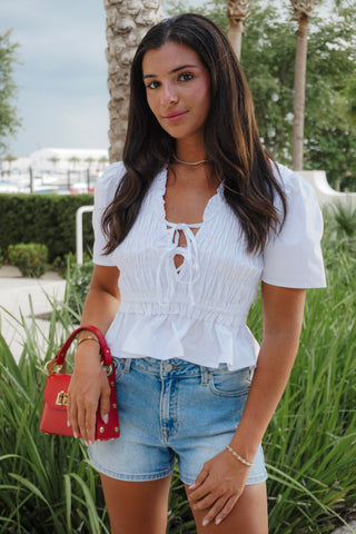 Woman wearing a white blouse and denim shorts with a red handbag outdoors.