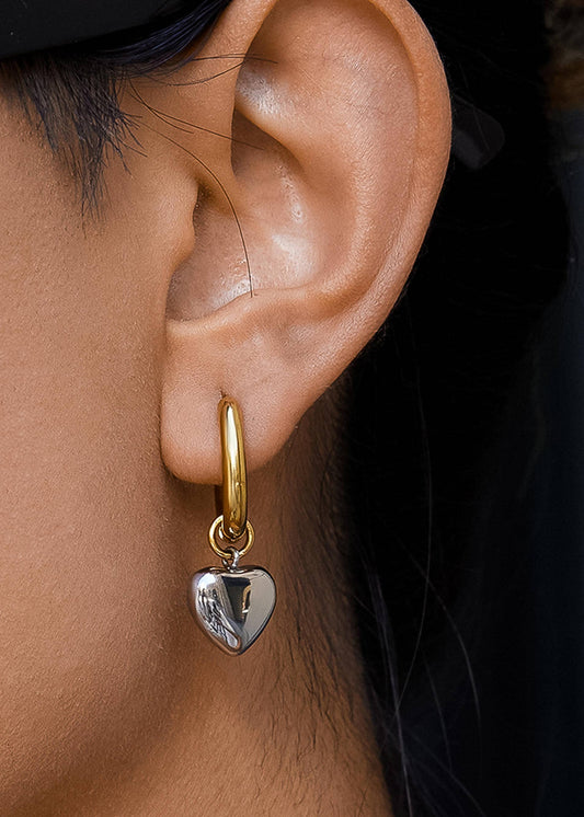 Gold hoop earring with a heart-shaped pendant worn by a person.