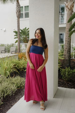 A woman wearing a two-tone maxi dress with a navy blue strapless bodice and a vibrant magenta pink skirt, standing outdoors on a tiled walkway next to a white building with palm trees.