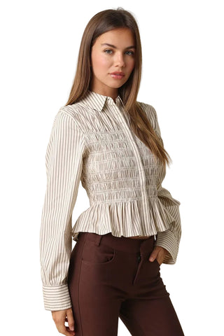 A woman modeling a long-sleeve collared blouse with vertical stripes and a smocked peplum waist, paired with dark brown pants, against a solid beige background.