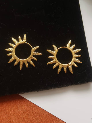 A pair of textured gold hoop earrings with sunburst details, displayed on a black velvet jewelry stand.