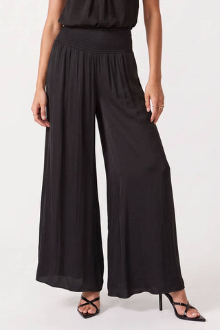 A model wearing black wide-leg pleated pants with a smocked waistband, paired with black strappy heels, shown from the waist down on a white studio background.