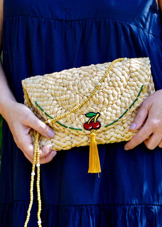Person holding a woven clutch with cherry design against a green leafy background