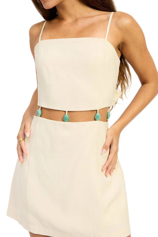 Woman wearing a beige dress with a belt featuring turquoise pom-poms on a white background