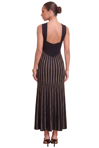 A woman shown from the back wearing a sleeveless black maxi dress with a structured open back and a pleated skirt with vertical metallic-looking stripes, against a plain beige studio background.