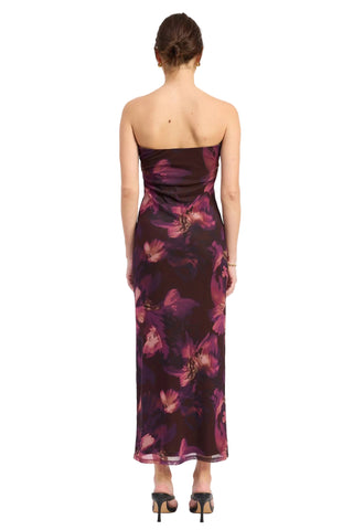 A woman viewed from the back wearing a strapless dark maxi dress with a purple and magenta floral print, paired with black high heels, against a plain white studio background.