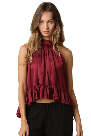 A woman modeling a sleeveless high-neck top in deep burgundy with a gathered ruffle hem, paired with black pants, against a solid beige background.
